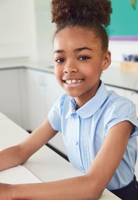 Lyseblå bluse med korte ærmer, rund krave og dekorative syninger. Sidder ved et skrivebord med en notesbog og smiler til kameraet.