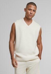 Pulover tricotat crem, fără mâneci, cu decolteu în V și tiv cu model texturat, realizat dintr-un material moale. Modelul stă pe un fundal gri.