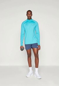 Turquoise langemouwen sporttop met rits, marineblauwe shorts, witte sneakers, houdt een dumbbell vast, gladde textuur, sportief ontwerp.