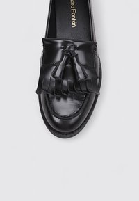 Mocasines de cuero negro con un acabado brillante, que presentan un detalle de borlas con flecos en la parte frontal y costuras en contraste a lo largo de la puntera.