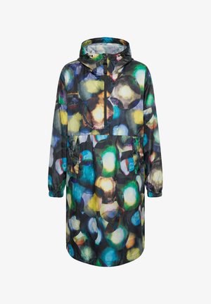 Femme portant un imperméable coloré à motif abstrait, les mains dans les poches, jean bleu et bottines bleu clair, debout sur fond blanc.