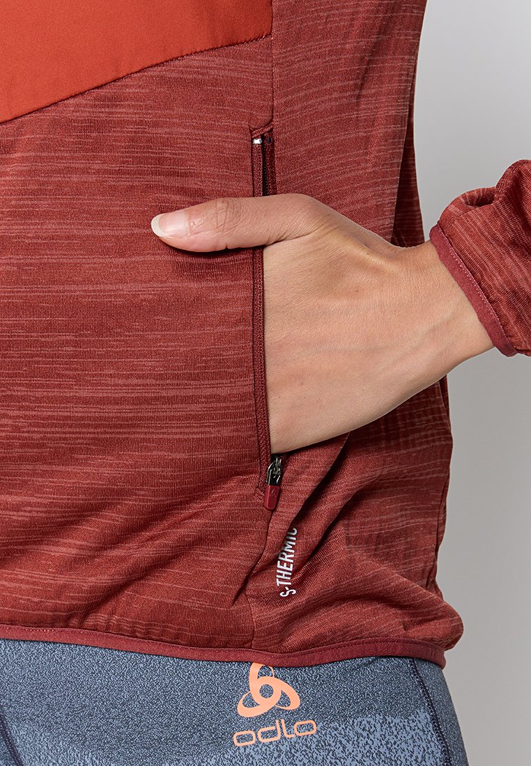 Rotes, strukturiertes Sportoberteil mit Reißverschlusstasche, feuchtigkeitsableitendem Stoff und kontrastierendem oberen Abschnitt. Logo sichtbar in der Nähe des Saums.