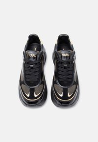 Μαύρα και χρυσά αθλητικά παπούτσια με άνω μέρος από δίχτυ και σουέτ, με επενδεδυμένη γλώσσα, μαύρα κορδόνια και χοντρή μαύρη σόλα. Το λογότυπο εμφανίζεται στις πλευρές.