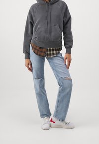 Felpa grigia con tasca frontale, indossata sopra una camicia a scacchi marrone e nera, abbinata a jeans strappati blu chiaro e sneakers bianche.