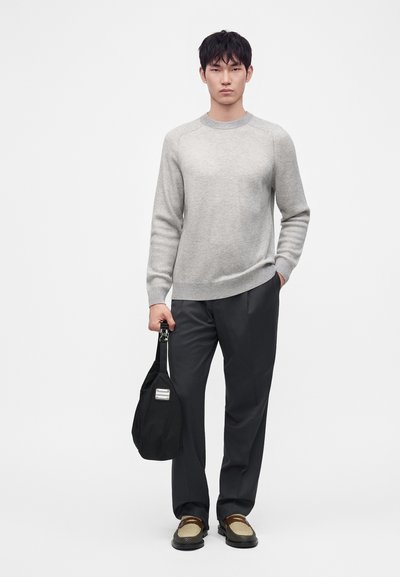 Jeune homme portant un pull gris clair, pantalon foncé, tenant un petit sac noir, debout sur un fond blanc uni.