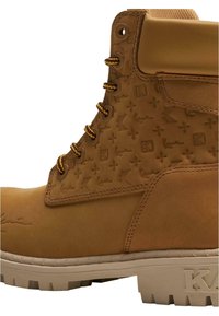 Brune lærstøvler med en struktureret design der har logoer og mønstre. Brune snørebånd, polstret krave, og en beige gummisål med godt greb.