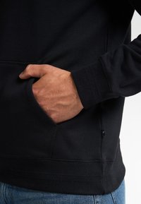 Černá mikina s kapucí vyrobená z měkké tkaniny, s klokaní kapsou, žebrovanými manžetami a lemem. Diskrétní štítek značky na boku.