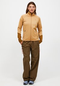 Giacca a zip tan con accenti marroni, due tasche con zip. Pantaloni marroni, larghi, con lacci rossi regolabili e più tasche.