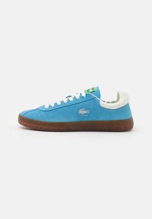 Blauwe lage sneaker van suede met witte veters, witte gevoerde kraag, bruine doorzichtige zool en Lacoste-logo aan de zijkant.