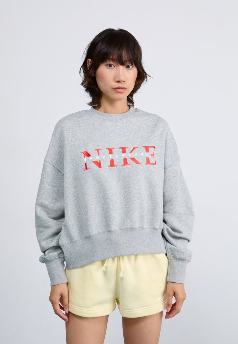 Sivi dukserica s crvenim "NIKE" logom, širokim rukavima i rebrastim završetkom. Uparena s žutim šortsevima, prikazujući opušteni kroj i mekanu teksturu.