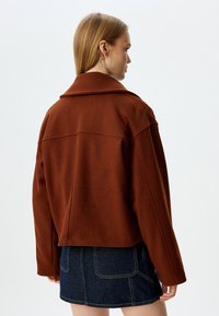 Giacca marrone in misto lana con colletto strutturato, design corto e cuciture in evidenza. Indossata sopra una gonna di jeans scura.