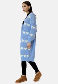 Cardigan lavorato a maglia di grandi dimensioni in azzurro chiaro con motivi a cuori bianchi, dotato di maniche lunghe e polsini a coste, abbinato a leggings neri e stivali color sabbia.