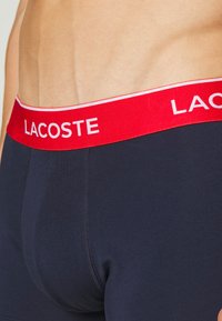 Boxer-briefs en coton bleu marine avec une ceinture élastique rouge portant le logo "LACOSTE" en blanc. Texture lisse et coupe ajustée.