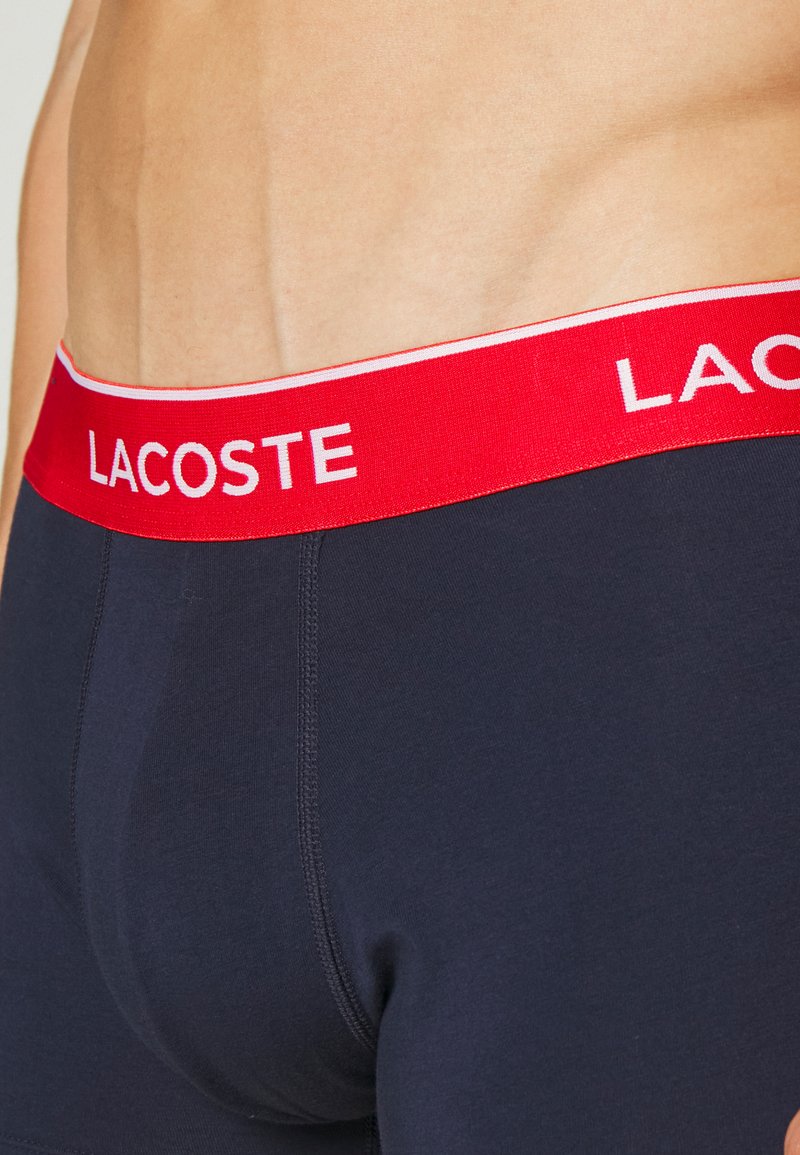 Boxer-briefs en coton bleu marine avec une ceinture élastique rouge portant le logo "LACOSTE" en blanc. Texture lisse et coupe ajustée.
