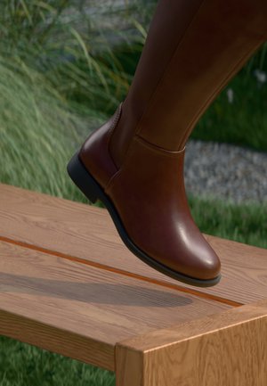 Brune skinn kne-høy støvel med rund tå og glatt tekstur, med en lav svart hæl og elastisk panel for fleksibilitet.