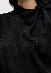 Blusa nera con motivo geometrico testurizzato, un collo alto e un grande dettaglio a fiocco sulla collar. Il tessuto è semi-trasparente.