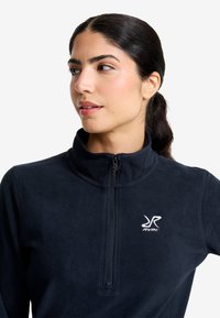 Frau mit dunklem Haar im Pferdeschwanz, die einen marineblauen Fleece-Pullover mit einem halben Reißverschluss und dem weißen RVRC-Logo auf der Brust trägt.
