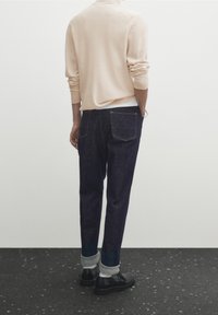 Man wearing light beige sweater, dark cuffed jeans, black shoes, standing with hands partially in pockets against plain background.