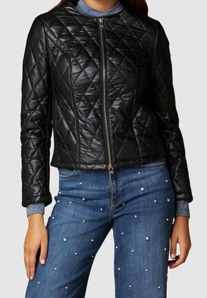 Žena v černé prošívané bundě na zip přes modré tričko s dlouhým rukávem, doplněná modrými džínami zdobenými malými bílými perlami.