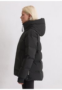 Frau mit kurzem blonden Haar, die eine schwarze Steppjacke mit hohem Kragen und Kapuze trägt, steht im Profil vor einer weißen Wand.