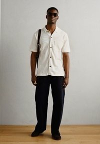 Camicetta a maniche corte color crema con un motivo texturizzato, bottoni neri, abbinata a pantaloni blu scuro e scarpe slip-on marroni. Accessorio: borsa a spalla marrone.