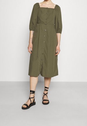 Robe olive, mi-longue, avec décolleté carré, manches bouffantes et fermeture à boutons à l'avant. Associée à des sandales à lanières noires.