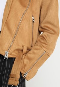 Veste en suède beige avec des zips en argent, une taille ceinturée et des poches zippées, avec une finition légèrement texturée et douce.