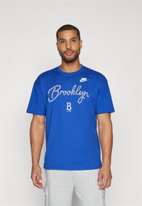Mėlyna medvilninė trumpa rankovė marškinėliai su užrašu "Brooklyn" baltu kursyvu, Nike logotipas kairėje krūtinės pusėje, apvalios kaklo iškirptės dizainas.