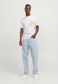 Hvid kortærmet t-shirt med en gul mønstret lomme, parret med lyseblå jeans og hvide sneakers, stående mod en neutral baggrund.