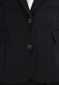 Musta räätälöity bleiseri, jossa on kaksi nappia, terävät kaulukset ja sivutaskut. Valmistettu sileästä kankaasta, joka istuu hyvin.