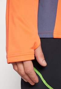 T-shirt de sport à manches longues, en tissu orange vif avec une texture, panneau en contraste bleu marine et surpiqûres vertes le long de l' ourlet.