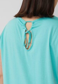 Een turquoise, kortärmige top met een ronde halslijn heeft een keyhole uitsnijding aan de achterkant, vastgezet met strikken. Zachte, gladde stof structuur.