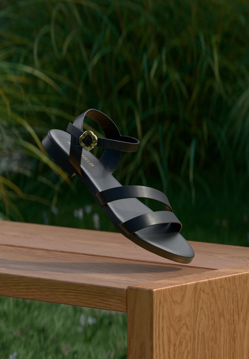 Sandale en cuir noir avec trois larges brides, bride de cheville avec accent doré, semelle plate, présentée sur un fond vert naturel.