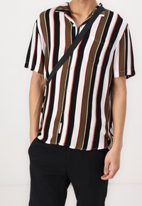 Mies, joka on pukeutunut lyhythihaiseseen nappipaitaan, jossa on pystysuorat mustat, valkoiset, oliivinvihreät ja punaiset raidat, mustiin housuihin ja mustaan ristikassi-hihnaan.