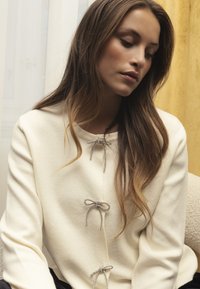Cream-colored sweater with a soft texture, featuring three jeweled bow accents down the front. Round neckline and long sleeves.