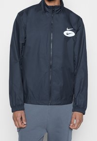 Marineblauwe windbreaker jas met een hoge kraag, elastische manchetten, ritssluiting aan de voorkant en een wit Nike-logo op de linkerkant van de borst. Gladde, lichte stof.