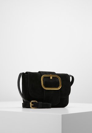 Sac bandoulière en daim noir avec grande boucle rectangulaire dorée et sangle réglable, posé sur un socle blanc sur fond uni.