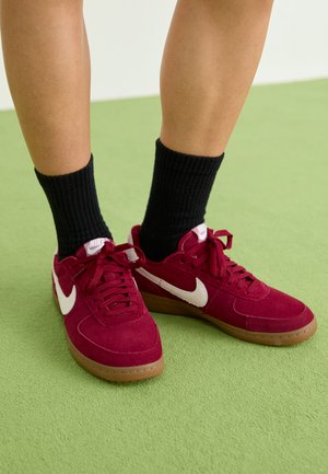 Piros velúr sportcipők fehér Nike pipa logóval, kerek orral, gumitalppal és azonos piros fűzőkkel. Fekete bordázott zoknival viselve. Zöld háttér.
