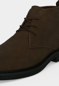 Bottine en daim marron avec semelle noire et lacets marron foncé noués en un simple motif croisé.