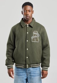 Olive green varsity jacket with a large embroidered letter 'A', featuring a collared neck, white snap buttons, and striped cuffs and hem.