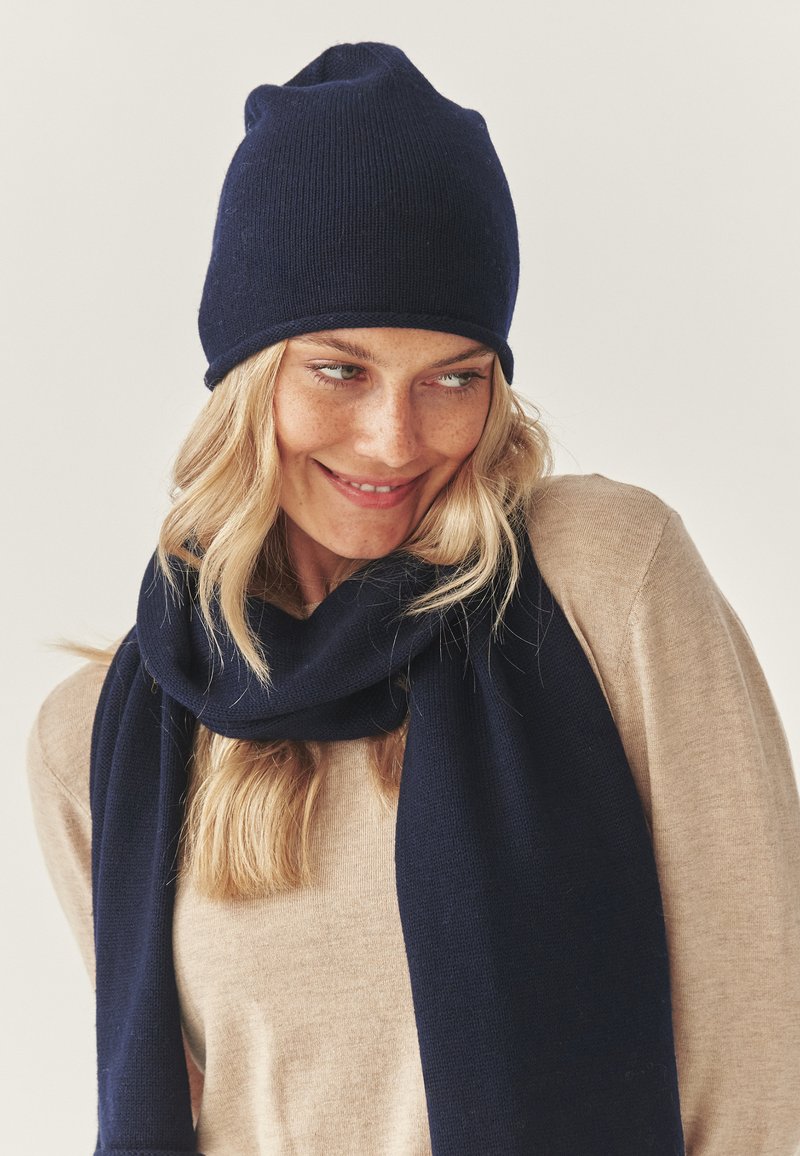 Mornarska plava pletena kapa s laganim visećim rubom, uparena s odgovarajućim šalicom od istog materijala, nošena preko bež dugih rukava.