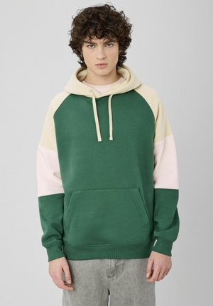 Sweat à capuche avec des blocs de couleurs vert, crème et rose. Comprend une poche avant, des cordons de serrage, ainsi qu'un ourlet et des poignets côtelés. Texture de tissu douce.