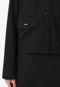 Veste noire cropped en tissu lisse, avec des boutons dorés en accent et deux poches poitrine avec rabats.