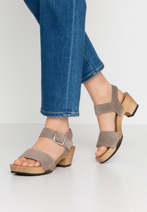 Persoon die blauwe spijkerbroek en beige suède open-toe sandalen met houten blokhakken en enkelbandjes draagt, staat op een witte vloer.