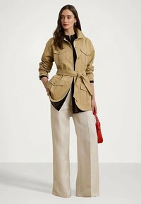 Ženska nosi bež jakno s pasom in velikimi žepi, smetana široke hlače do tal, drži majhno rdečo torbico, stoji ob enostavni ozadju.