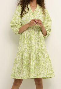 Weißes Kleid mit grünem abstraktem Muster, V-Ausschnitt, Ballonärmeln und einem gestaffelten Rockdesign. Der Stoff wirkt strukturiert.