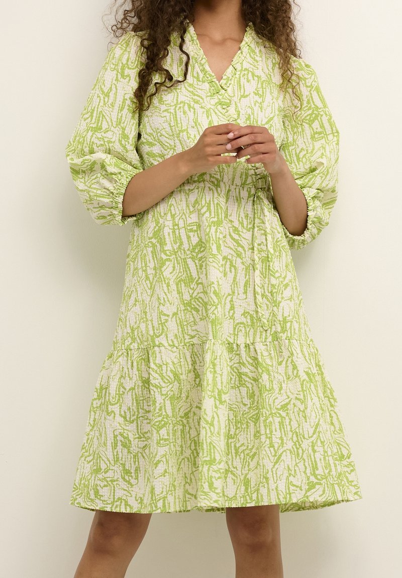 Weißes Kleid mit grünem abstraktem Muster, V-Ausschnitt, Ballonärmeln und einem gestaffelten Rockdesign. Der Stoff wirkt strukturiert.