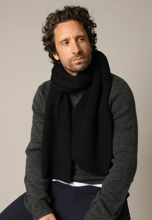 Écharpe noire côtelée drapée autour du cou sur un pull gris texturé. Les matériaux semblent doux et chauds, adaptés pour porter par temps froid.