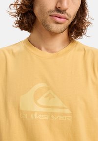 Žluté bavlněné tričko s velkým, reliéfním logem Quiksilver v světlé barvě, klasickým kulatým výstřihem a krátkými rukávy.