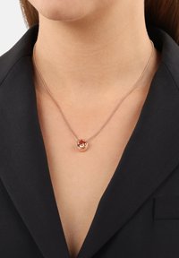 Roségoldene Halskette mit einem runden, orangefarbenen Edelsteinhänger, an einer feinen Kette, die gegen ein schwarzes Blazer getragen wird. Strukturiertes, glattes Finish.